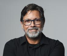Homem sorrindo, de cabelo curto, liso e castanho escuro, barba grisalha, usando óculos de grau de armação retangular azul naval, e camisa preta. Ao fundo, parede cinza.